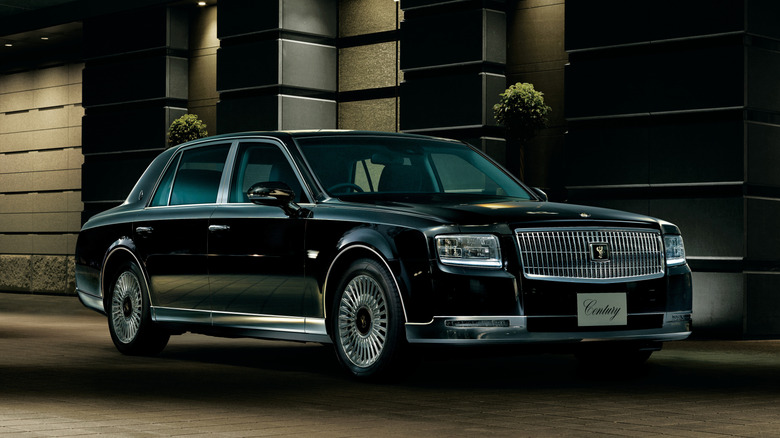 A black Toyota Century sedan parked in front of a modern building, showcasing its classic boxy luxury profile.