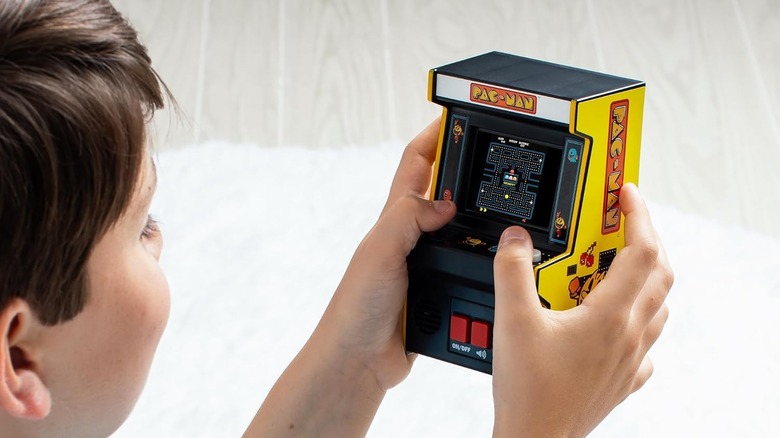 A close-up of a child playing a handheld Mini Pac-Man arcade cabinet with a red joystick.