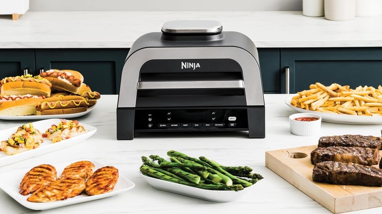 A Ninja Foodi 6-in-1 indoor grill and multi-cooker on a kitchen counter with cooked food around it.