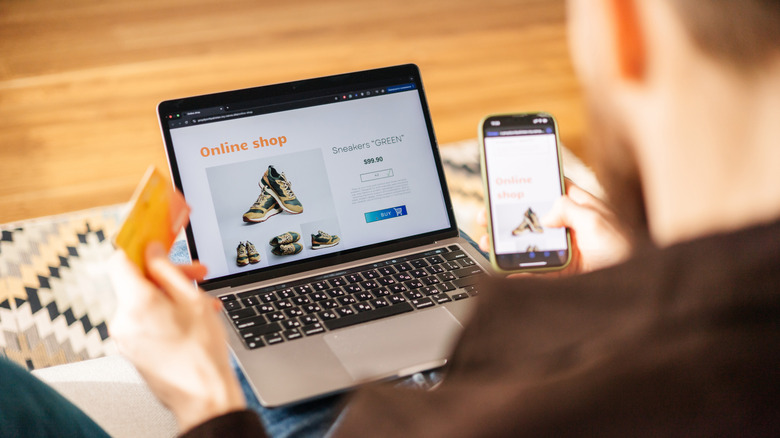 A man looking at shoes on a laptop while holding a smartphone.