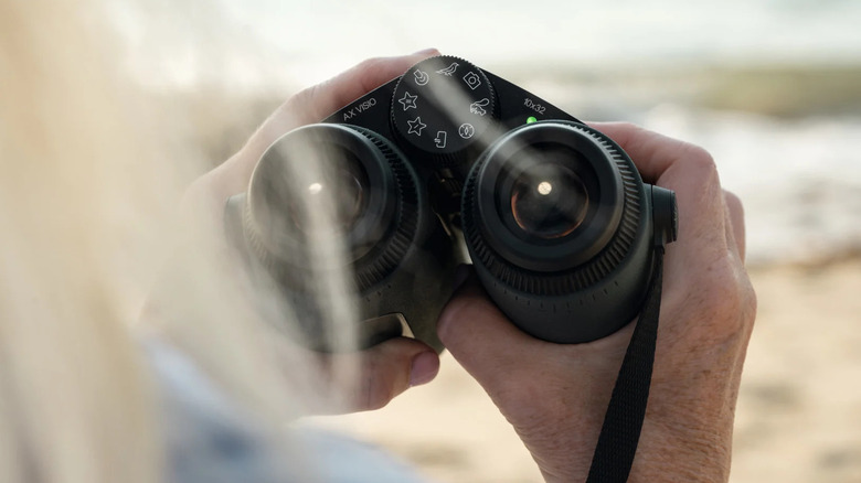 A person holding a pair of Swarovski AX Visio smart binoculars in an outdoor, natural setting.