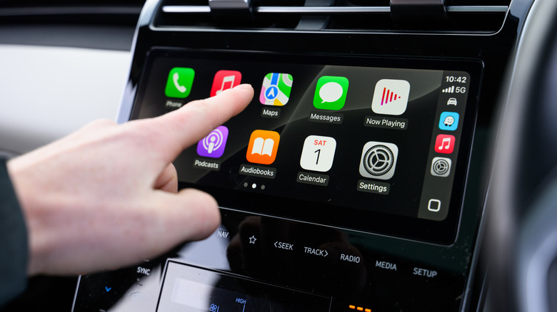 A hand tapping the touchscreen of a car's head unit to navigate Apple CarPlay.