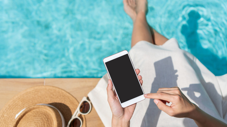 A person sitting cross-legged by a swimming pool holding their smartphone.