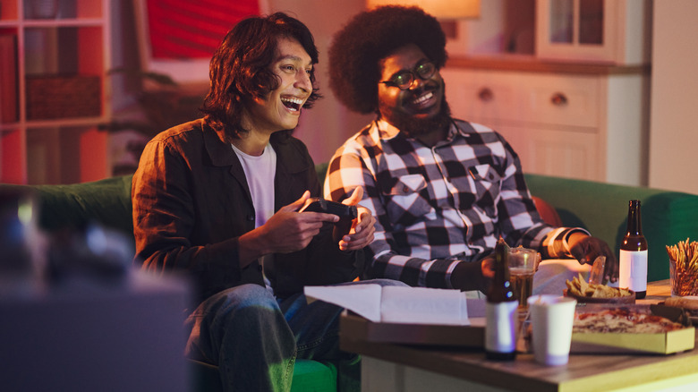 Two people playing video games with controllers in a setting with snacks and food on the table.