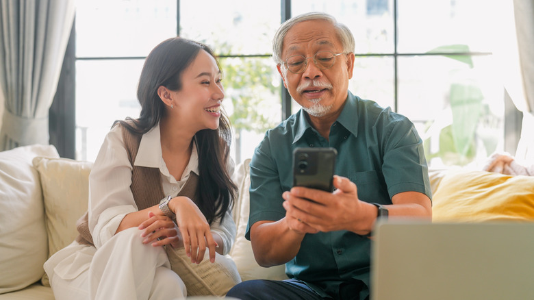 An older man sitting with a younger woman as they both look at and enjoy a smartphone screen together.