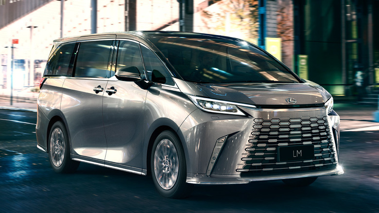 A silver Lexus LM luxury minivan driving on a city road at night surrounded by streetlights.