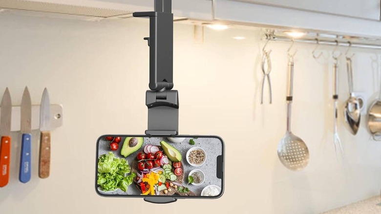 A close-up of the Perilogics phone mount clamped onto a household shelf, holding a smartphone horizontally.