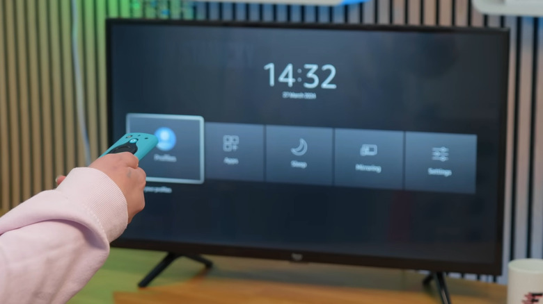 A close-up of a hand holding an Alexa Voice Remote pointing at a TV screen showing a settings shortcut.