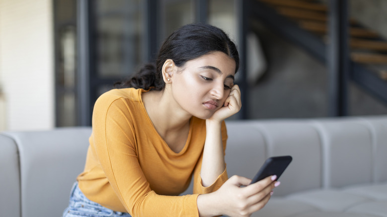 A woman sitting on a couch looking at her phone with a bored and disinterested expression.
