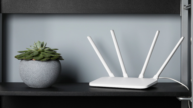 A white Wi-Fi router positioned on a high black shelf next to a decorative plant.