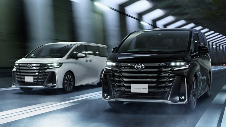 A black and a white Toyota Vellfire driving through a brightly lit tunnel, showcasing their bold front grilles.